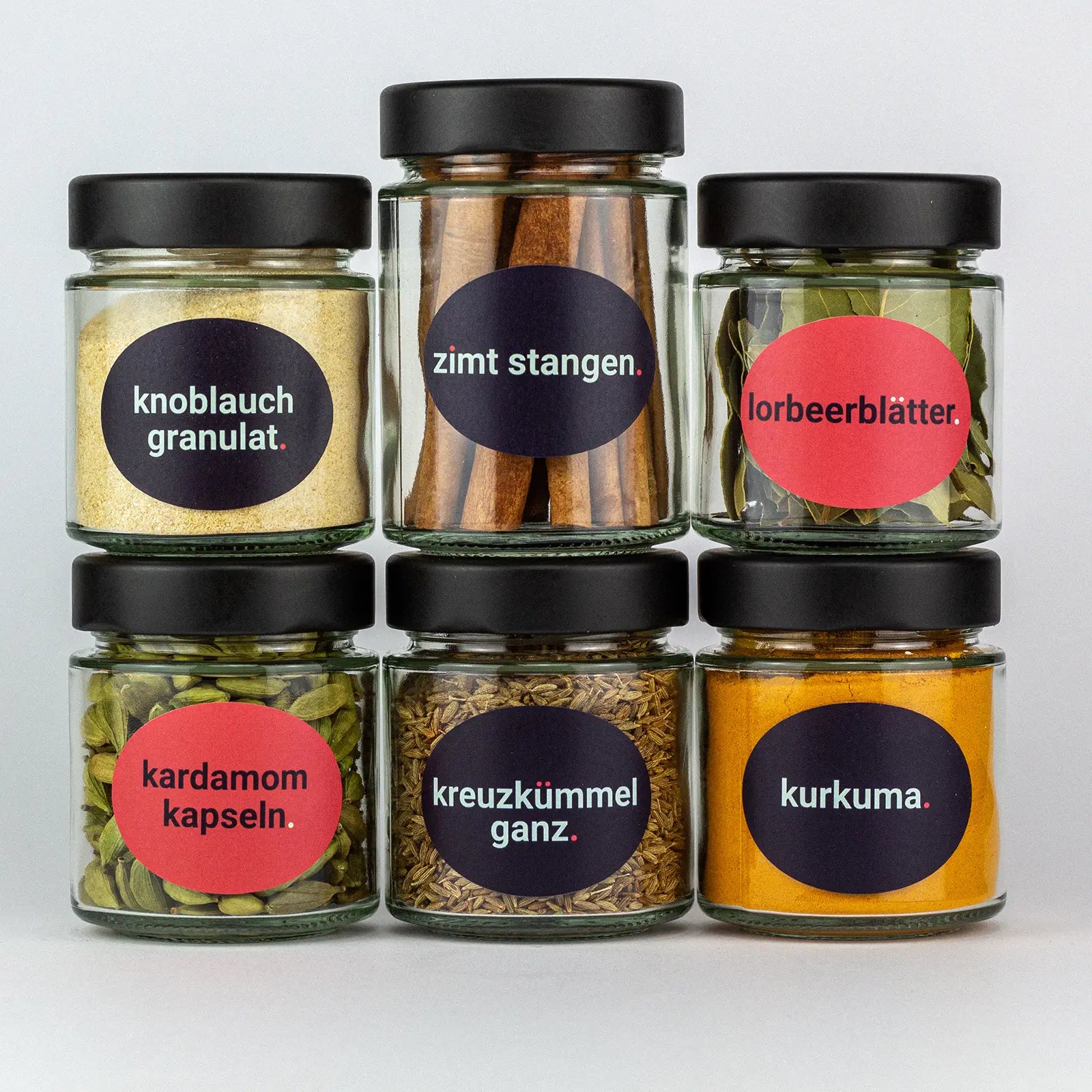 Die Gewürze des Reingewürze Set Knoblauchgranulat, Zimtstangen, Lorbeerblätter, Kardamomkapseln, Kreuzkümel Ganz und Kurkuma in Gläsern aufeinandergestapelt.