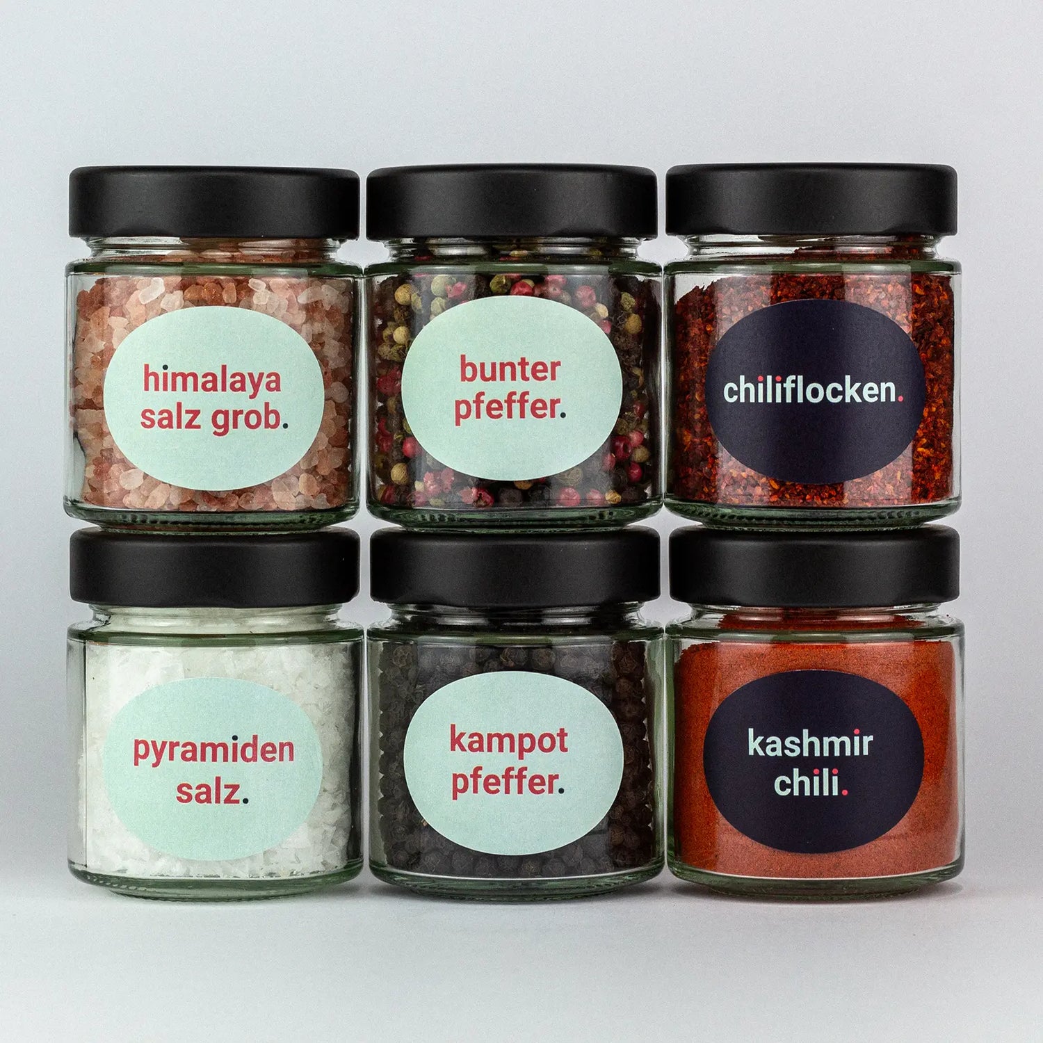 Die Gewürze des Salz & Pfeffer & Chili Set Himalaya Salz Grob, Bunter Pfeffer, Chiliflocken, Pyramidensalz, Kampotpfeffer und Kashmirchili in Gläsern aufeinandergestapelt.